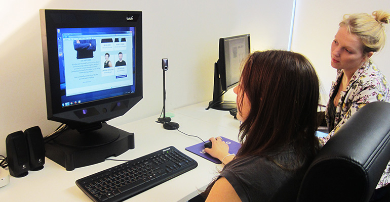 Image of two women in front of a computer. One is navigating through a website using a mouse. The other woman is leaning in and looking at the screen